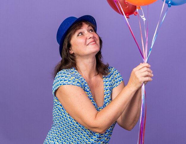 Felice e allegra donna di mezza età con cappello da festa che tiene un mazzo di palloncini colorati guardandoli sorridenti che celebrano ampiamente la festa di compleanno in piedi sul muro viola