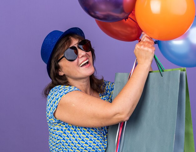 Felice e allegra donna di mezza età con cappello da festa e occhiali che tengono un mazzo di palloncini colorati e sacchetti di carta con regali sorridenti che celebrano la festa di compleanno in piedi sul muro viola