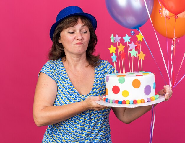 Felice e allegra donna di mezza età in cappello da festa con palloncini colorati che tengono la torta di compleanno guardandola con un sorriso sul viso che celebra la festa di compleanno in piedi sul muro rosa