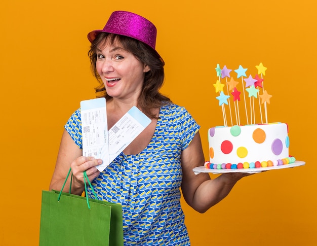 Felice e allegra donna di mezza età in cappello del partito che tiene il sacchetto di carta con i regali che tengono la torta di compleanno e i biglietti aerei