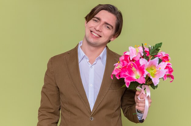 Felice e allegro giovane azienda bouquet di fiori guardando davanti sorridendo allegramente andando a congratularsi con la giornata internazionale della donna in piedi sopra la parete verde