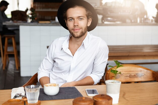 Felice e allegro giovane uomo barbuto in copricapo alla moda che mangia caffè, seduto al tavolo di legno nell'interno del caffè moderno, aspettando la sua ragazza, progettando di proporle questa giornata di sole