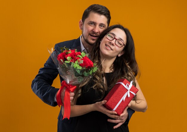 Felice e bella coppia uomo con bouquet di rose e donna con regalo che abbraccia felice innamorato festeggiando San Valentino sul muro arancione over