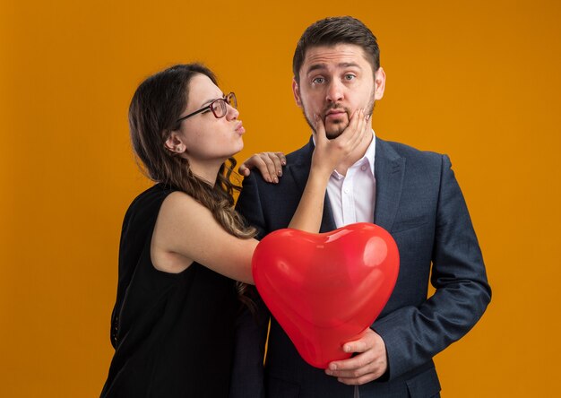 Felice e bella coppia uomo e donna con palloncino rosso a forma di cuore felice innamorato che celebra il giorno di san valentino sul muro arancione