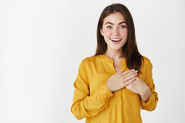 Felice e felice giovane donna che guarda con ammirazione e gioia, tenendosi per mano sul cuore in segno di apprezzamento per il dono, in piedi toccato sul muro bianco.