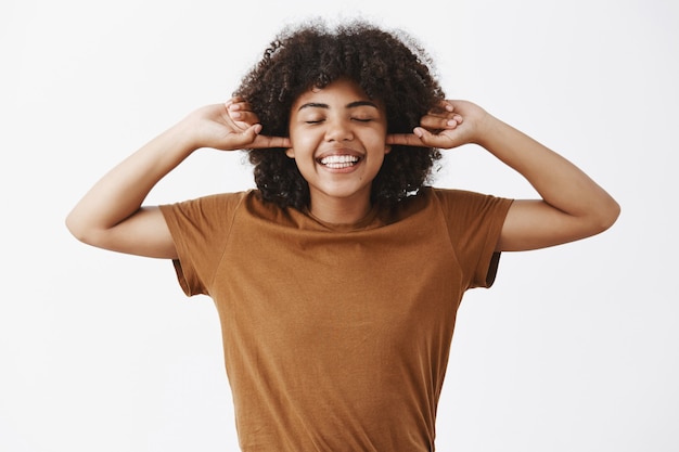 Felice e gioiosa donna afroamericana emotiva con acconciatura afro in t-shirt marrone alla moda che copre le orecchie con le dita indice sorridente ampiamente e chiudendo gli occhi godendo il silenzio