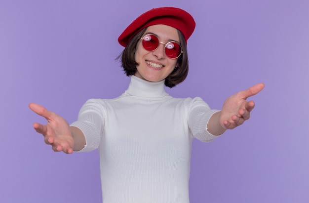 felice e positiva giovane donna con i capelli corti in dolcevita bianco che indossa berretto e occhiali da sole rossi
