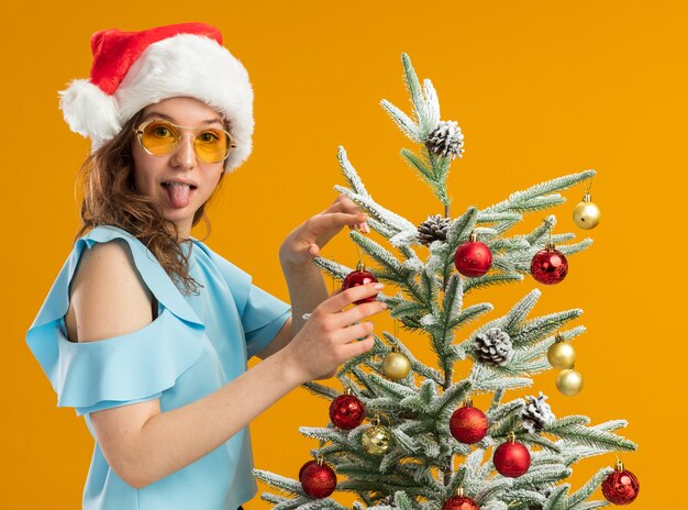 Felice e positivo giovane donna nella parte superiore blu e santa hat indossando occhiali gialli decorare albero di natale spuntavano lingua guardando la fotocamera in piedi su sfondo arancione