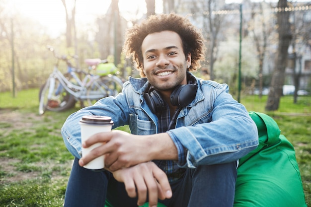 Felice e positivo giovane studente maschio con afro-acconciatura in abiti alla moda seduto nel parco mentre sorride ampiamente e beve caffè.