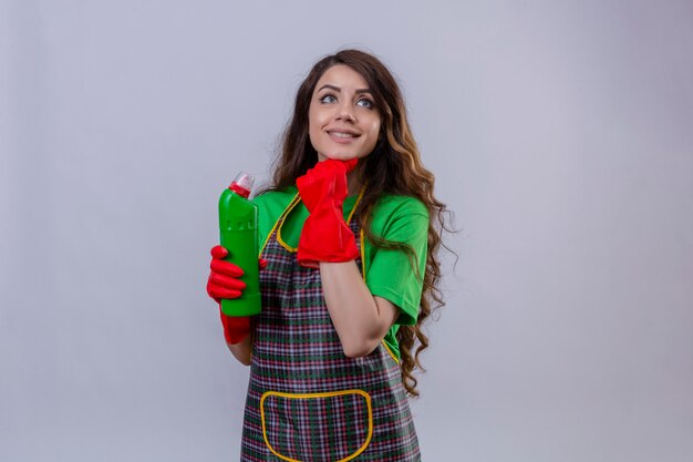 Felice e premurosa giovane bella donna con lunghi capelli ondulati che indossa grembiule e guanti di gomma in piedi con la mano sul mento mentre si tiene una bottiglia di prodotti per la pulizia che guarda lontano in piedi