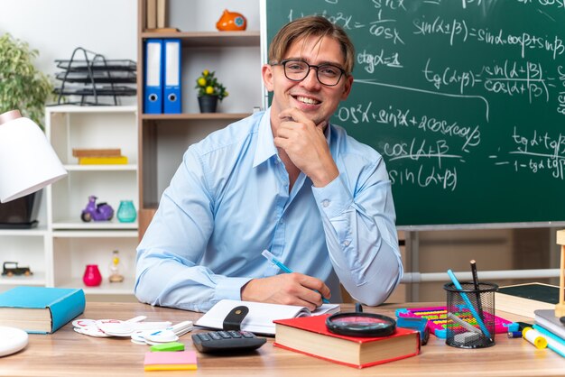 Felice e sorridente giovane insegnante maschio seduto al banco di scuola con libri penna e note davanti alla lavagna in classe in