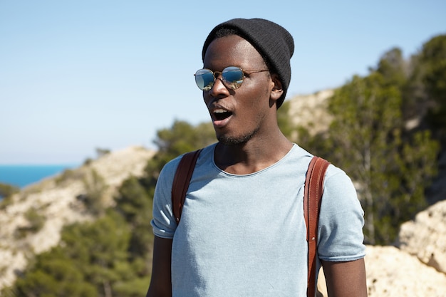 Felice eccitato giovane viaggiatore maschio afroamericano in eleganti tonalità e copricapo che sembrano sbalorditi, tenendo la bocca spalancata mentre in piedi in alto nelle montagne e ammirando la splendida vista sottostante