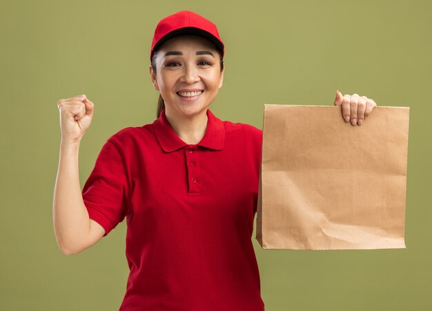 Felice ed eccitata giovane donna delle consegne in uniforme rossa e berretto che tiene in mano un pacchetto di carta con un sorriso sul viso che stringe il pugno in piedi sul muro verde