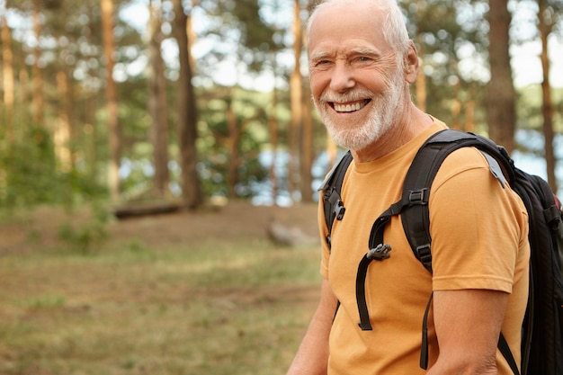 Felice energico uomo in pensione con zaino nero dietro la schiena che sorride ampiamente, godendosi le escursioni nei boschi nella soleggiata giornata autunnale. Colpo all'aperto dell'uomo anziano con la barba che cammina nella foresta