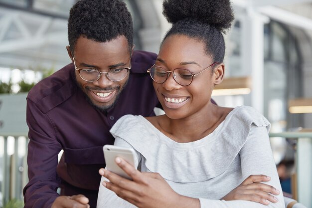 Felice femmina africana in occhiali tipi messaggio sul moderno smart phone mentre il suo ragazzo sta vicino a lei e guarda lo schermo