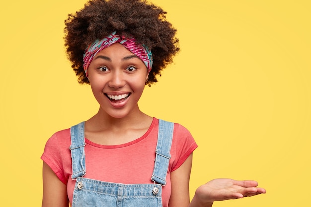 Felice femmina afroamericana con un sorriso amichevole, mostra i denti bianchi, vestita con maglietta casual e tuta, tiene il palmo sollevato, mostra uno spazio vuoto sul muro giallo. Persone e concetto di etnia