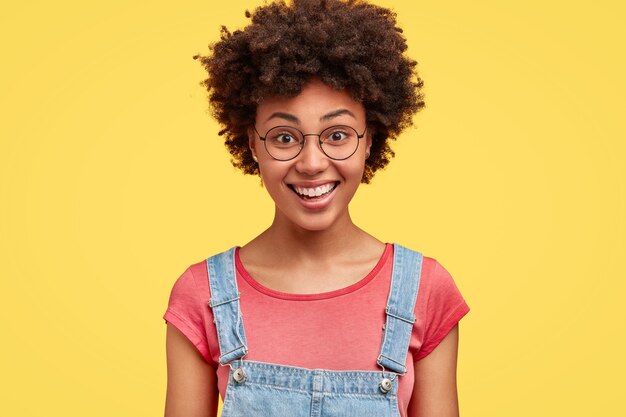 Felice femmina dalla pelle scura con acconciatura afro, ha un'espressione soddisfatta, mostra denti bianchi perfetti, è di buon umore dopo l'appuntamento con il fidanzato, isolata sopra il muro giallo. Concetto di emozioni