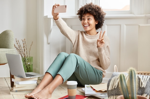 Felice femmina dalla pelle scura fa la videochiamata con il cellulare, mostra il segno di pace