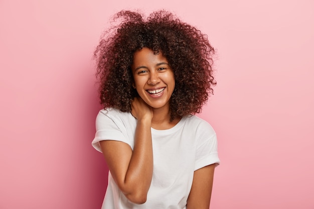 Felice femmina dalla pelle scura ha una conversazione divertente, guarda con un ampio sorriso, intrattenuta da un amico, ha un'espressione del viso felicissima, vestita con abiti di tutti i giorni, sta al coperto. Concetto di emozioni