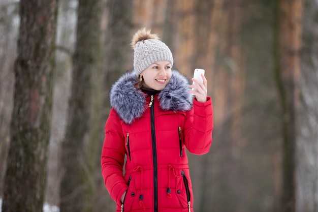 Felice femmina sorridente in giacca invernale rossa guarda sul suo cellulare, all&#39;aperto