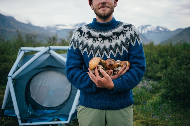 Felice, fiero uomo raccoglitore in tradizionale maglione di lana blu con ornamenti si trova su un campeggio in montagna, tiene in braccio un mucchio di funghi deliziosi e biologici
