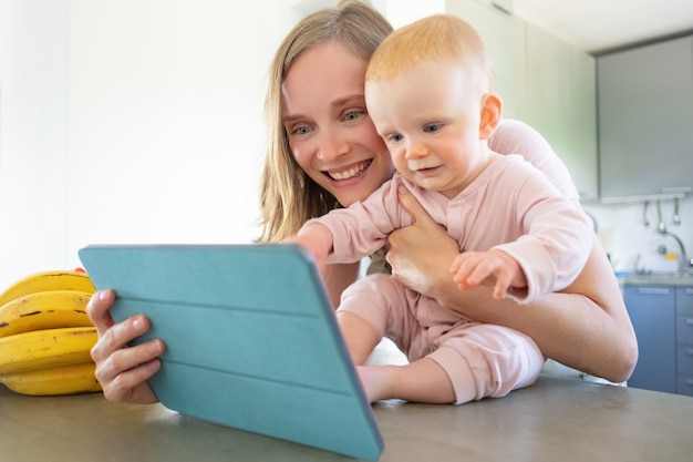 Felice gioiosa mamma e figlia bambino parlando alla famiglia in cucina, utilizzando tablet per videochiamata, sorridendo insieme allo schermo. Assistenza all'infanzia o concetto di comunicazione online