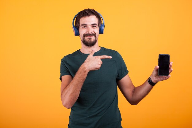 Felice gioioso uomo che indossa le cuffie e indica il suo smartphone isolato su sfondo arancione in studio.