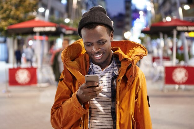 Felice giovane afro-americano vestito elegantemente in cappotto invernale e cappello avendo sera a piedi da solo per le strade della città straniera, gli amici di messaggistica sul gadget elettronico. Persone e tecnologia moderna