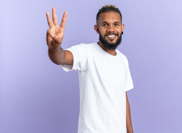 Felice giovane afroamericano in maglietta bianca che guarda la telecamera che mostra e punta verso l'alto con le dita numero tre sorridenti