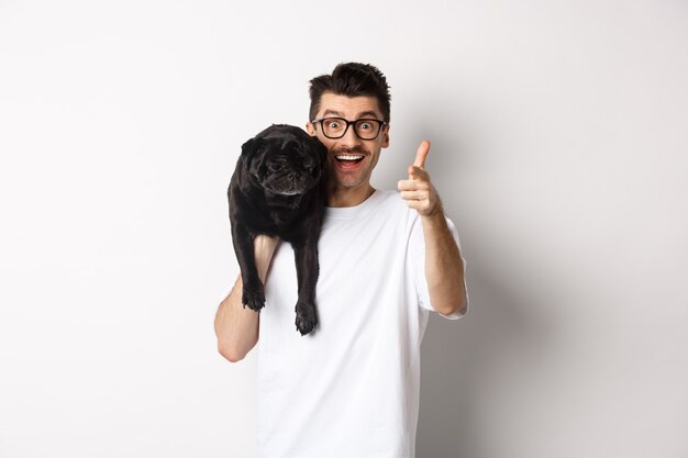 Felice giovane azienda simpatico cane nero sulla spalla e che punta alla telecamera. Il ragazzo hipster porta il carlino sulla spalla e fissa la telecamera eccitato, in piedi su sfondo bianco.