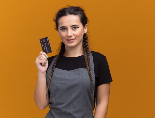 Felice giovane barbiere femminile in uniforme con carta di credito isolata sulla parete arancione