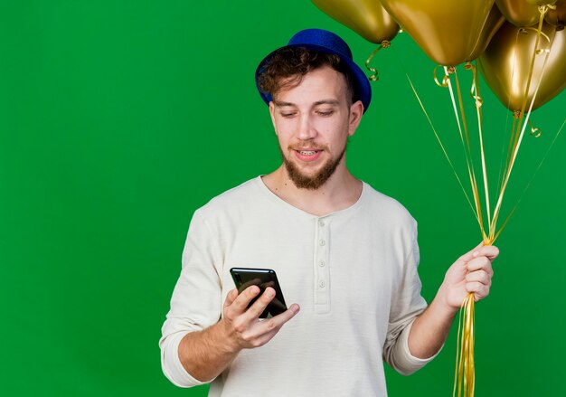 Felice giovane bel partito slavo ragazzo che indossa il cappello del partito che tiene palloncini e telefono cellulare guardando il telefono isolato su sfondo verde con spazio di copia