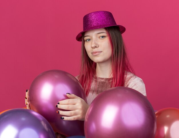 Felice giovane bella donna che indossa un cappello da festa in piedi dietro palloncini isolati su una parete rosa