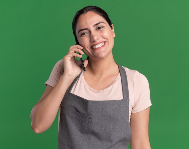 Felice giovane bella donna parrucchiere in grembiule guardando davanti sorridente mentre parla al telefono cellulare in piedi sopra la parete verde