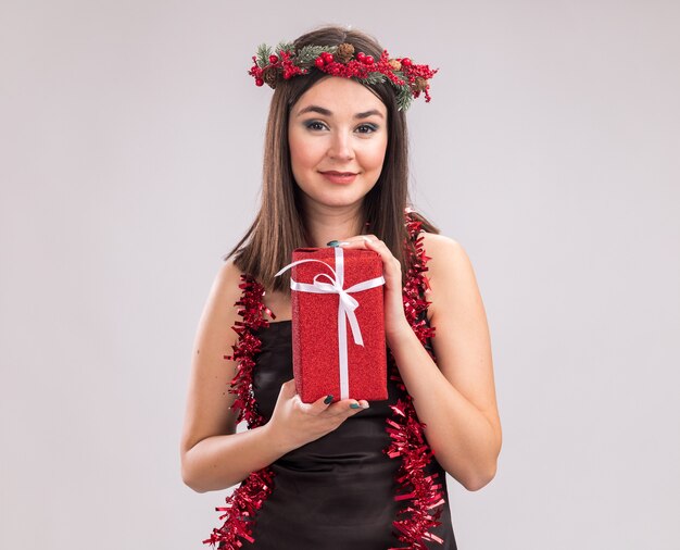 Felice giovane bella ragazza caucasica che indossa la testa di Natale ghirlanda e ghirlanda di orpelli intorno al collo tenendo il pacchetto regalo guardando la telecamera isolata su sfondo bianco con spazio di copia