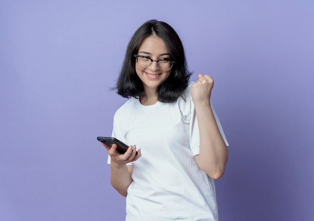 Felice giovane bella ragazza caucasica con gli occhiali guardando il lato tenendo il telefono cellulare e il pugno di serraggio isolato su sfondo viola con spazio di copia