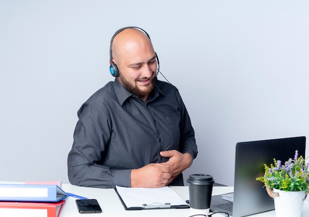 Felice giovane calvo call center uomo che indossa auricolare seduto alla scrivania con strumenti di lavoro guardando il computer portatile mettendo la mano sulla pancia isolato su sfondo bianco