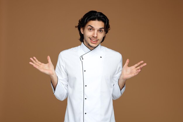 felice giovane chef maschio che indossa l'uniforme guardando la fotocamera che mostra le mani vuote isolate su sfondo marrone