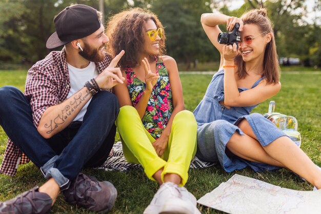 Felice giovane compagnia di amici seduti al parco, uomini e donne che si divertono insieme, colorato stile moda hipster estate, viaggiare con la fotocamera, scattare foto