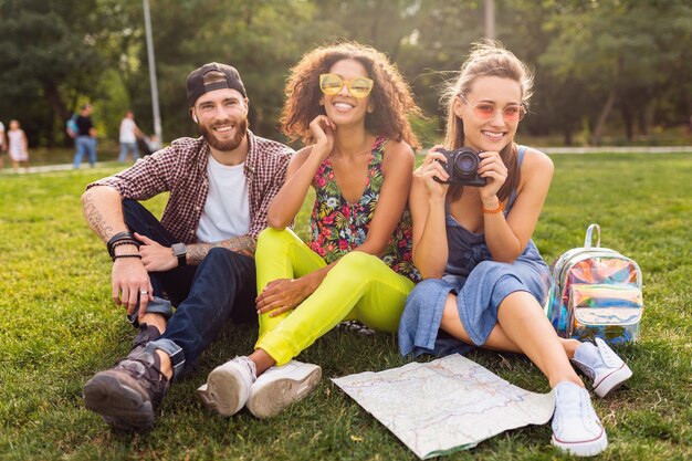 Felice giovane compagnia di amici seduti al parco, uomini e donne che si divertono insieme, viaggiano con la macchina fotografica, parlano, sorridono