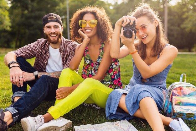 Felice giovane compagnia di amici seduti al parco, uomini e donne che si divertono insieme, viaggiano con la macchina fotografica, parlano, sorridono