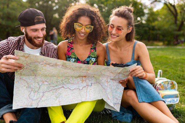 Felice giovane compagnia di amici seduti nel parco che viaggiano guardando nella mappa visite turistiche, uomini e donne che si divertono insieme