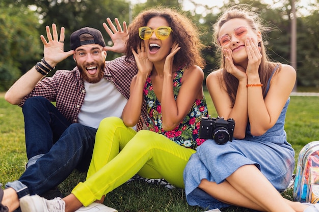 Felice giovane compagnia di amici seduti nel parco scherzare con facce buffe pazze, uomini e donne che si divertono insieme, viaggiando con la macchina fotografica