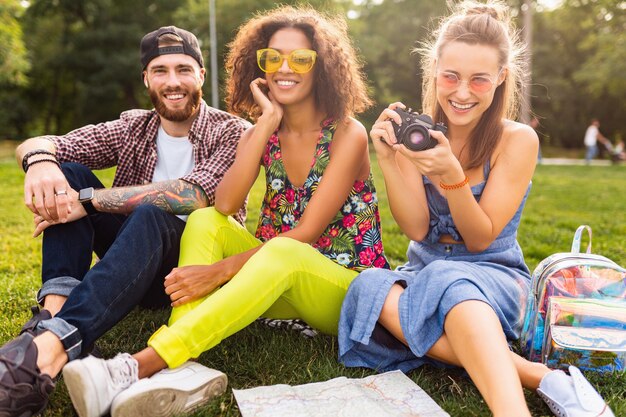 Felice giovane compagnia di amici seduti nel parco, uomini e donne che si divertono insieme, viaggiando con la macchina fotografica, ridendo schietti