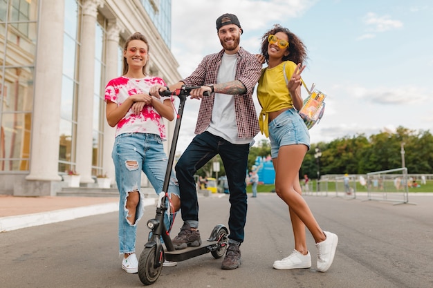 Felice giovane compagnia di amici sorridenti che camminano in strada con scooter elettrico, uomini e donne che si divertono insieme