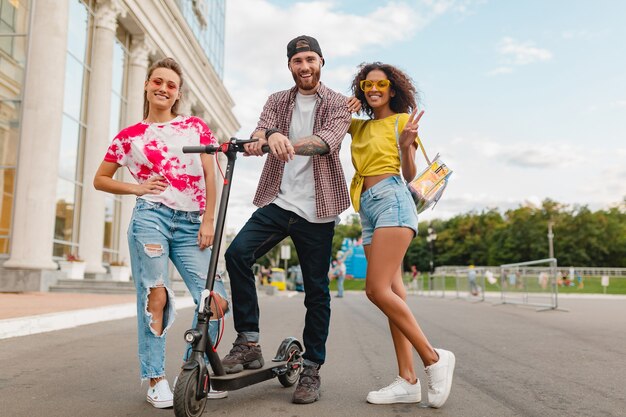 Felice giovane compagnia di amici sorridenti che camminano in strada con scooter elettrico, uomini e donne che si divertono insieme