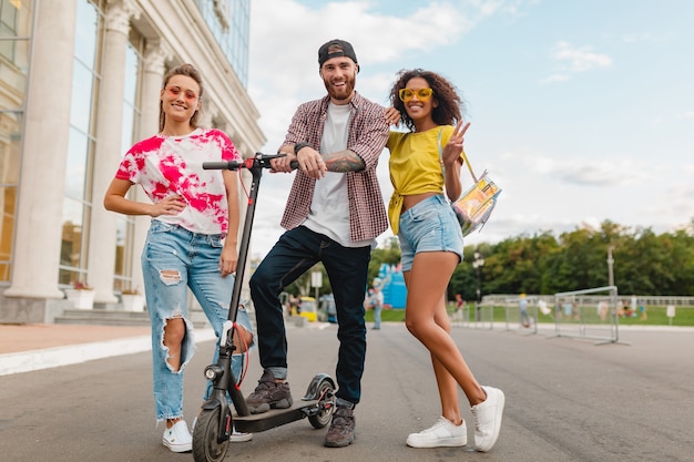 Felice giovane compagnia di amici sorridenti che camminano in strada con scooter elettrico, uomini e donne che si divertono insieme
