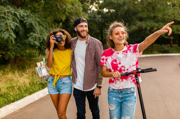 Felice giovane compagnia di amici sorridenti che camminano nel parco con scooter elettrico, uomini e donne che si divertono insieme, stile di moda hipster colorato estivo, viaggiando con la macchina fotografica, parlando, sorridendo