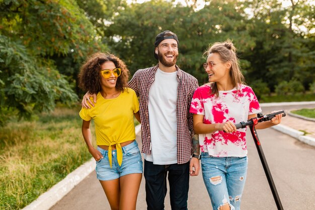 Felice giovane compagnia di amici sorridenti che camminano nel parco con scooter elettrico, uomini e donne che si divertono insieme