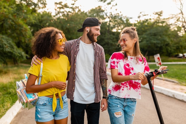 Felice giovane compagnia di amici sorridenti che camminano nel parco con scooter elettrico, uomini e donne che si divertono insieme